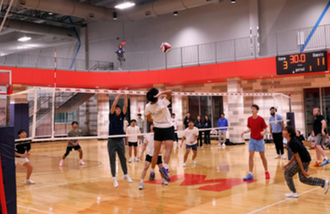 Two teams playing indoor volleyball, one player is in the air about to hit the ball over the net