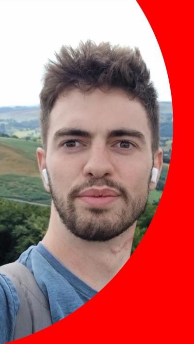 William Chapman has brown hair, a beard and moustache. He is wearing a blue T-shirt.