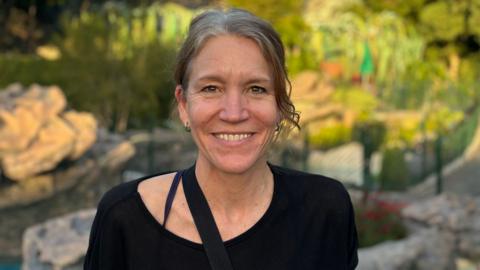 A woman pictured outside, looking directly at the camera and smiling. She has long brown/grey hair which is tied back. She is wearing a black top.