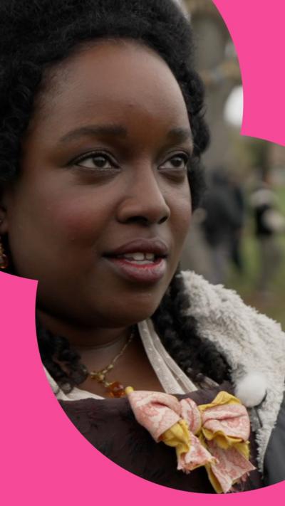 A woman has afro hair done in a luxurious style. She is dressed as an old-fashioned noblewoman, but wearing a modern coat with a fluffy lining over the top.