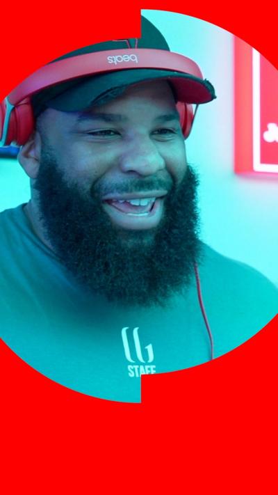 A DJ with a bushy black beard is talking to a reporter. He is wearing a black baseball cap and red headphones.