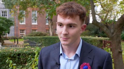 A young man in a blue suit with a rosette in a garden in London