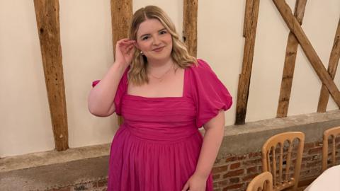 Jess is wearing a bright pink dress at a formal event. She has blonde wavy hair