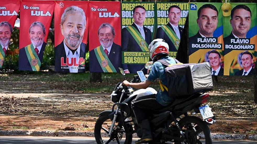 Flags and towels bearing the images of Brazil's President and presidential candidate Luiz Inacio Lula da Silva and right-wing senator and presidential candidate Flavio Bolsonaro are displayed for sale on a street in Brasilia as someone on a motorcycle drives by.