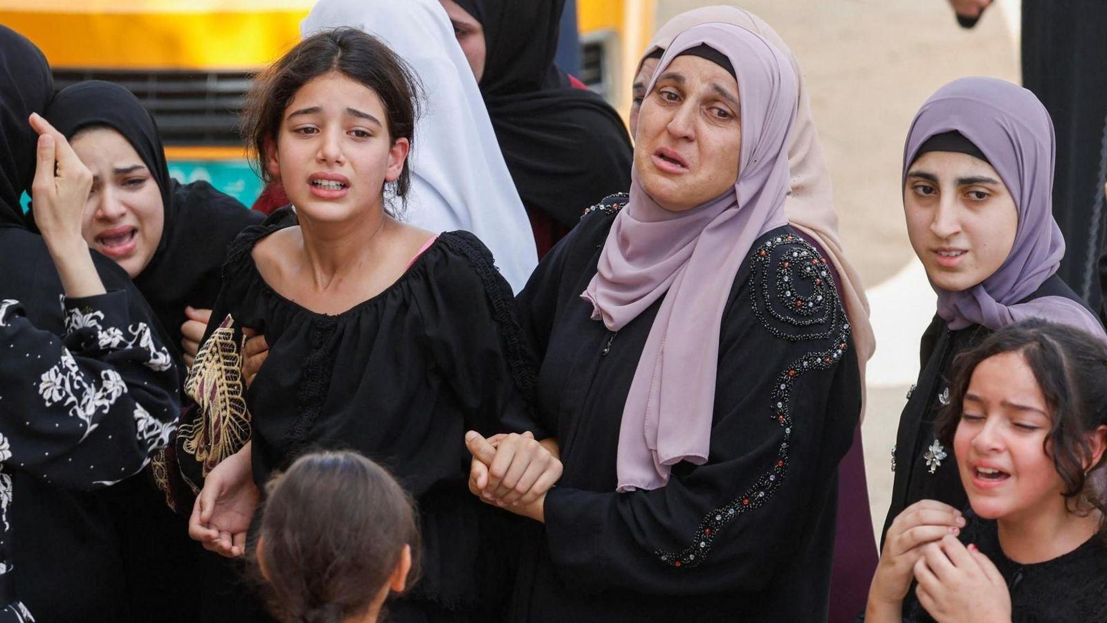 A woman wearing a black dress and purple headscarf holds the hand of a girl in a black top as a funeral. Three other girls are seen in black outfits, one wears a purple headscarf and one wears a black headscarf. They are all crying.