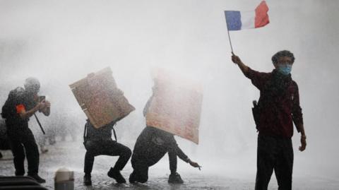 A masked protester holds a French flag as French police use water canon amid clashes during a demonstration by French high school and university students in Paris 