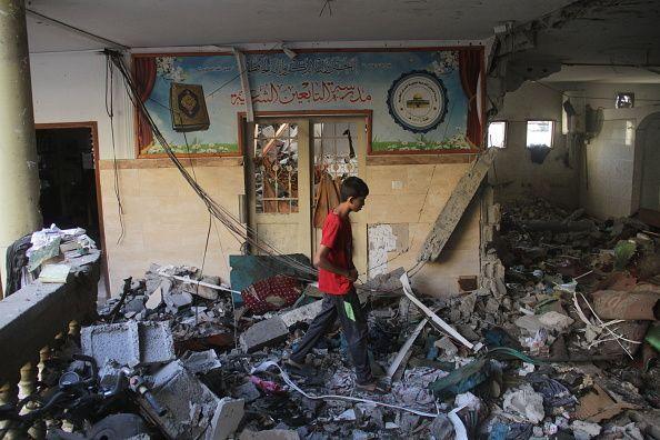 A boy in a red t-shirt walks through piles of rubble and strewn wires inside a school building. He is walking in front of a wall with a blue mural.