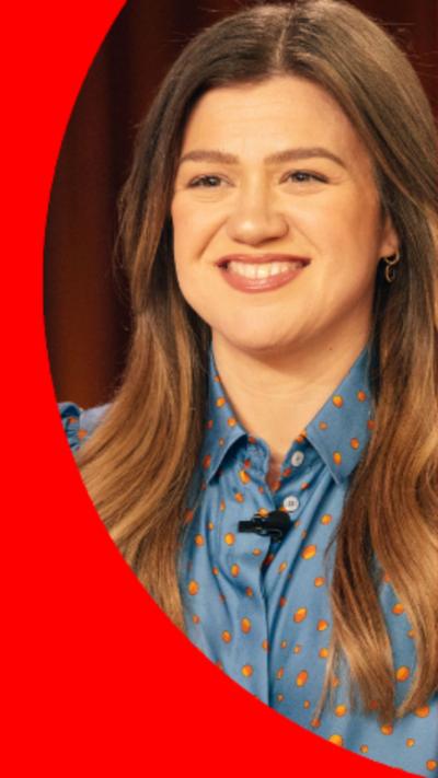 Kelly Clarkson has long brown hair and is wearing a blue shirt with yellow spots.
