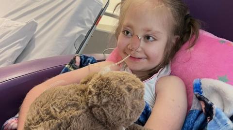 Willow sits in a chair in hospital cuddling a soft toy. She has a tube in her nose and is smiling