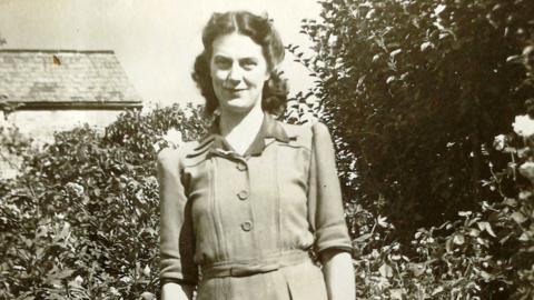 A black and white photo of a woman smiling at the camera in front of bushes