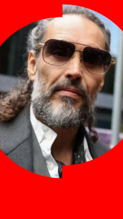 A close-up of Russell Brand with tied-back grey hair and a grey beard. He's wearing sunglasses.