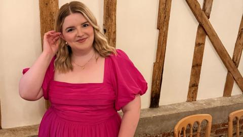 Jess is wearing a bright pink dress at a formal event. She has blonde wavy hair