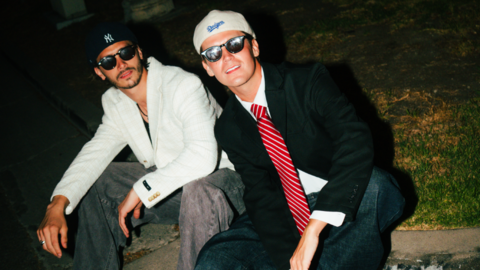 Two young men, sat next to each other on a pavement, both wearing baseball caps, sunglasses, jeans and blazers and looking at the camera. The one on the left has dark hair and facial hair and a white blazer and black t-shirt. The one on the right has a black blazer, white shirt and red striped tie and is clean shaven with short hair.
