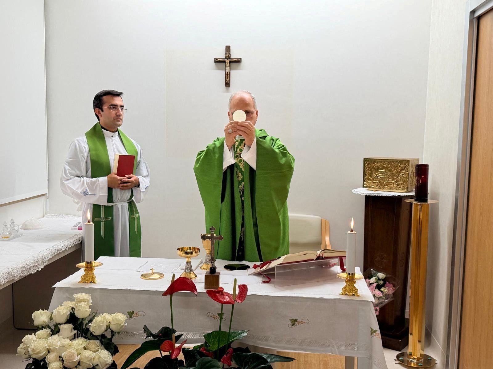 Pope Leo celebrates mass from hospital, he is wearing a green robe and standing before a white wall adorned with a wooden crucifix. He is standing next to his personal secretary who holds a red book and his wearing white and green. 