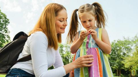 A little girl wearing a rainbow dress and looking down at a daisy chain in her hand. Her mother is crouching down next to her talking to her.