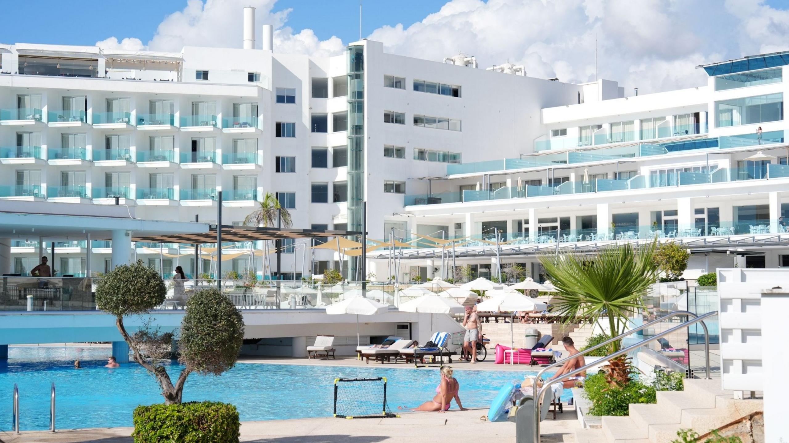 People lounging by a swimming pool at the hotel, which is a large white building with various balconies on its front.