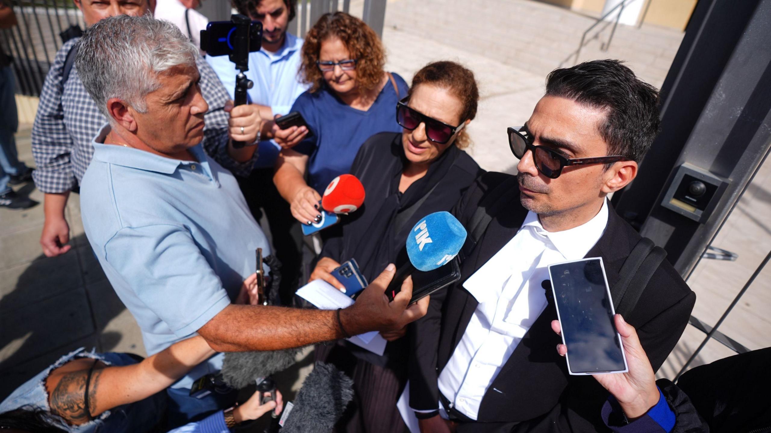 Alexandros Alexandrou, who is wearing a suit and black sunglasses, speaking to a crowd of reporters outside court.
