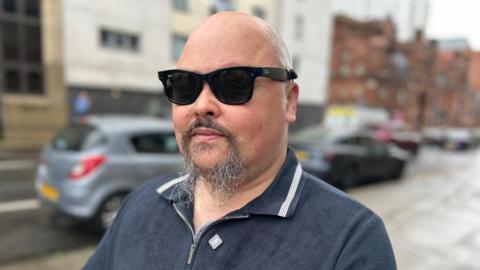 Portrait of Fraser Fleming wearing dark sunglasses and a navy zip-neck polo shirt, standing on a wet city street with parked cars and buildings blurred in the background.