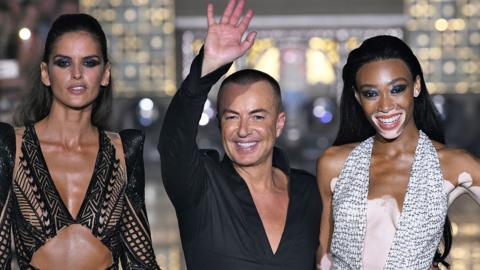 Two women and a man smiling down a runway. The man is waving.
