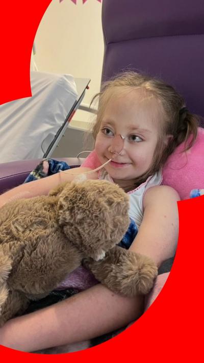 Willow sits in a chair in hospital cuddling a soft toy. She has a tube in her nose and is smiling