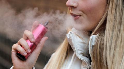 A blonde woman breaths out vapour as she holds a red vape in her hand