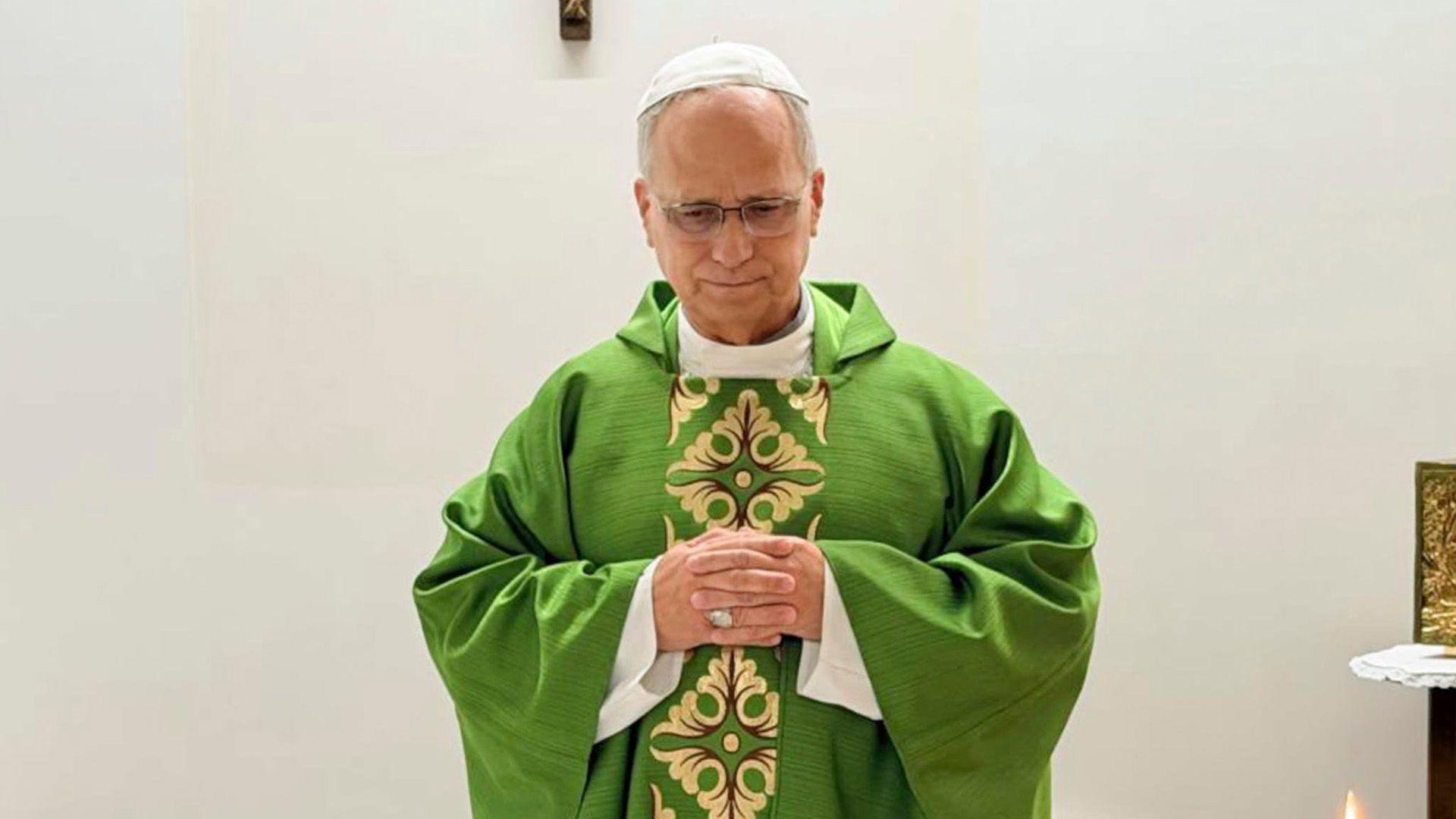 Pope Leo celebrates mass from hospital, wearing a green robe and standing before a white wall adorned with a wooden crucifix. 