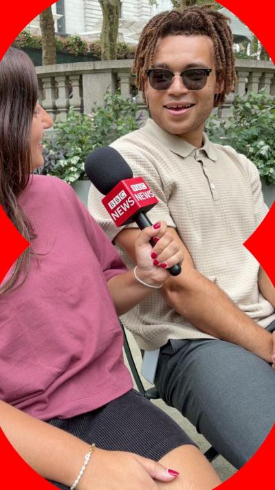 A woman holding a microphone talks to a man on a bench