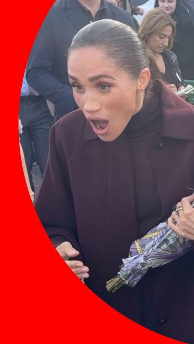 Meghan Markle is wearing a long burgundy coat. Her hair is gelled back into a ponytail. She has a surprised expression.