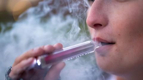 A cloud of vapour spreads around the partially obscured face of a woman holding a vape