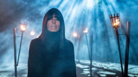 Claudia Winkleman in the Traitors. She is bathed in rays of moonlight poking through a forest canopy as braziers light the path behind her