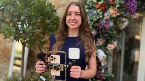 A young woman standing outdoors holding a smartphone video rig with a microphone and LED light attachment, in front of decorative greenery and a floral display. She is wearing a dark sleeveless jumpsuit and smiling at the camera.