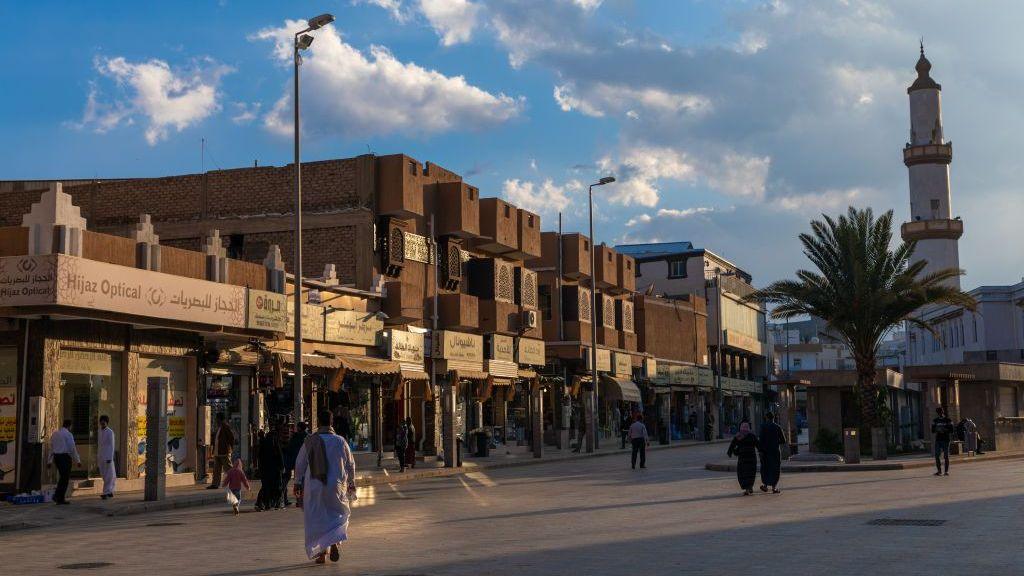 A broad street lined with low-rise shops stretches through a commercial district under late-afternoon light. Beige and brown buildings with Arabic signage face an open plaza where people walk along the paved road. A tall palm tree and a slender mosque minaret rise on the right side of the scene, while dramatic clouds fill the blue sky overhead.