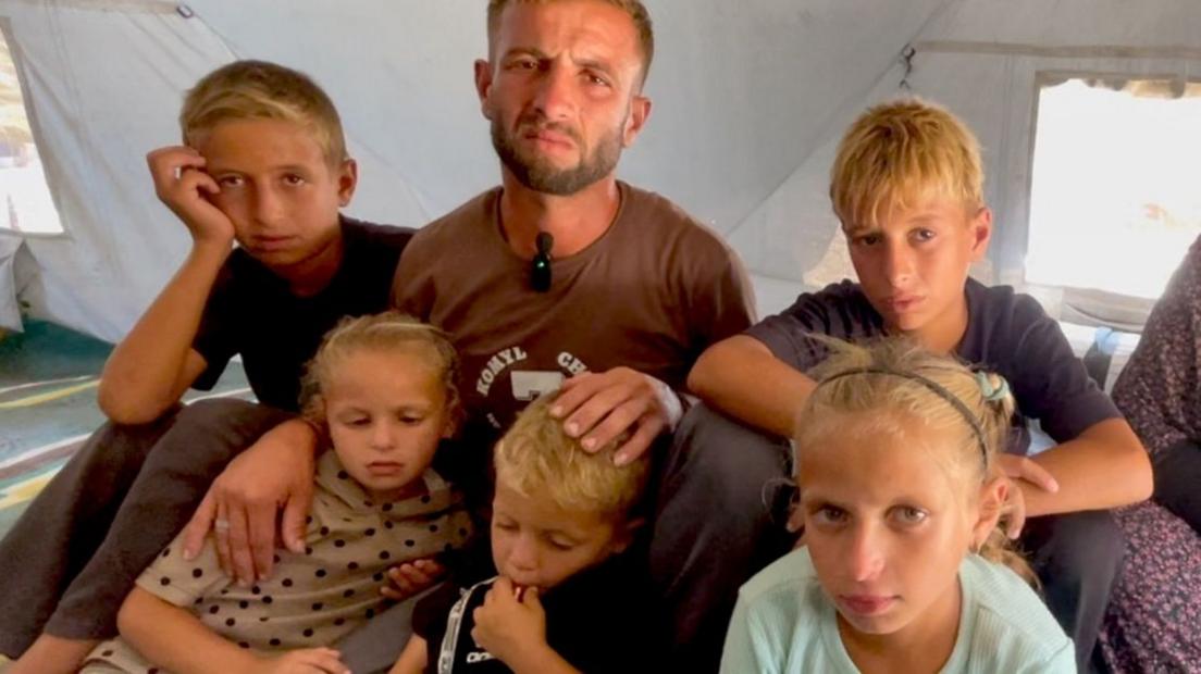 A man in his 30s sits with five children - two girls and three boys - inside a tent