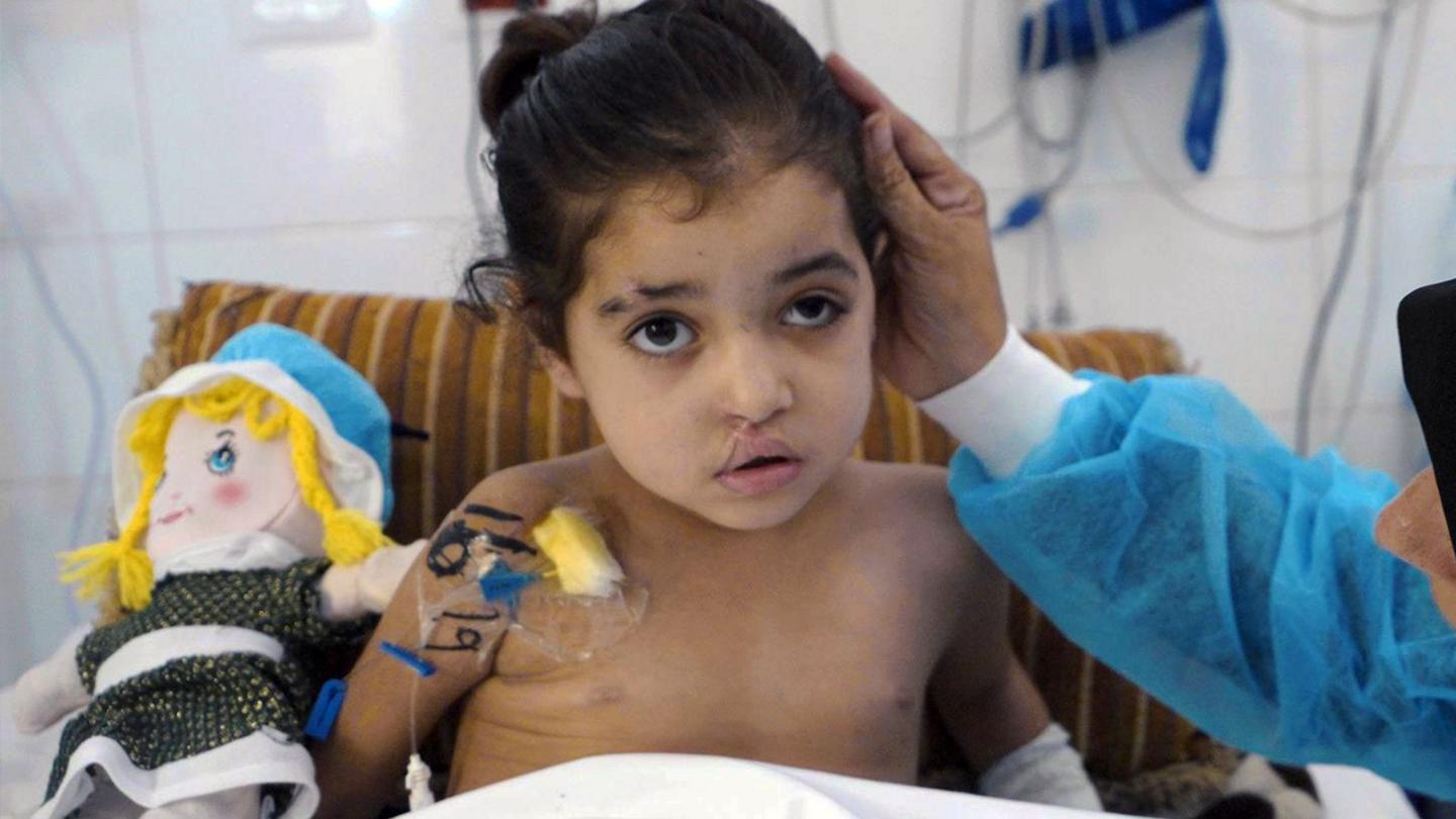 A young girl is seen in a hospital bed while a woman wearing scrubs strokes her hair
