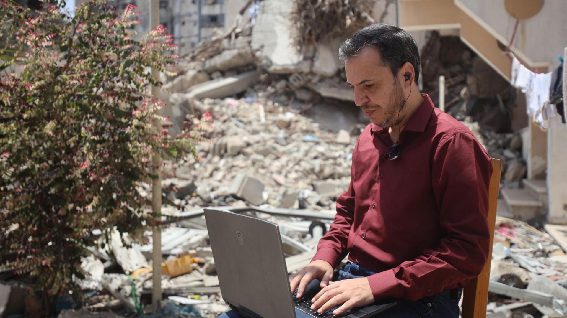A Palestinian man works on a laptop on a chair outside, surrounded by rubble, in Gaza 