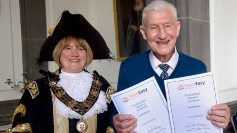 Alan is stood holding two Read Easy certificates next to a woman wearing a black hat. 