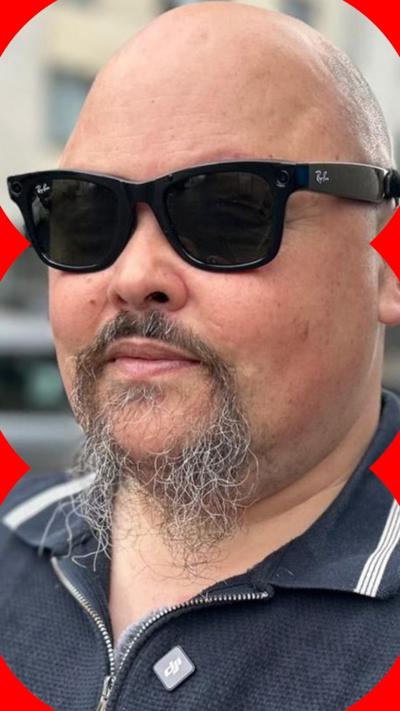 A man with a beard is wearing black 'smart glasses' with a small lens embedded in the frame. He is wearing a three-quarter zip navy jumper