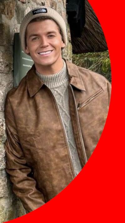 A young man wearing a beanie hat, grey jumper, brown jacket, brown chinos and white boots. He is leaning on the stone wall of a thatched building with a blue door and a sign that reads "muddy boots and paws welcome". He is smiling and looking at the camera.