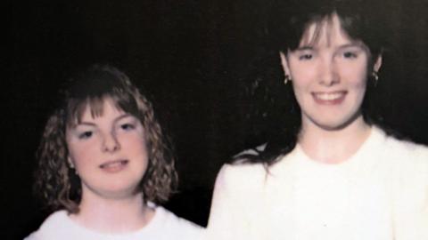 Vintage photograph of two young people standing side by side in light-coloured tops against a dark background. One has shoulder-length curly hair and the other has longer dark hair.