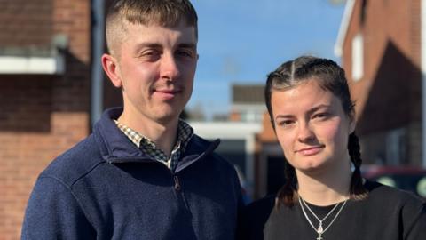 Ben and Meg stand outside. Ben is wearing a navy quarter zip top and checked shirt. Meg is wearing a black top and necklace. 