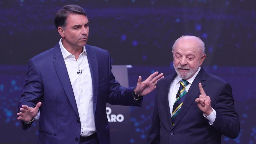 Luiz Inácio Lula da Silva gestures by holding up an index finger while Flávio Bolsonaro holds both his arms out in another gesture. Both are taking part in the presidential TV debate on 18 October. 