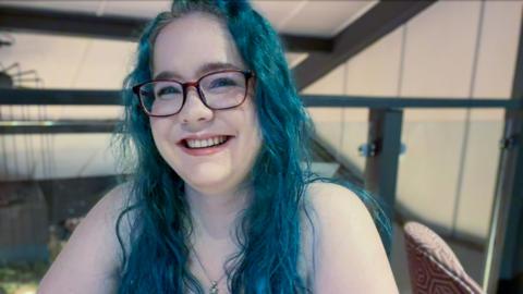 Olivia Miller-Davis, with long blue hair and glasses, sits smiling in a bar
