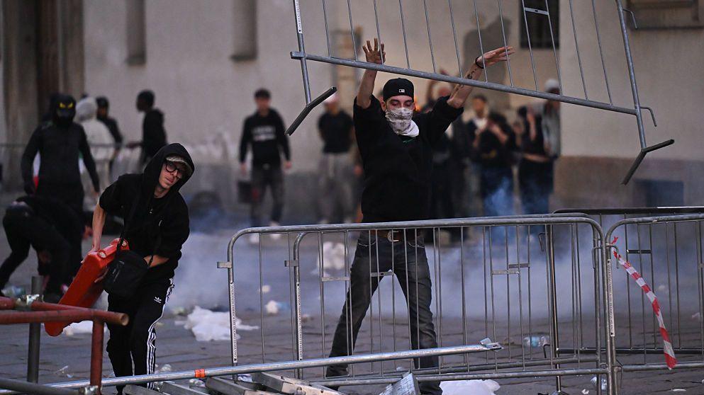 Man in black hoodie, black cap and white mask throwing a barrier up over another one while a man - also in a hoodie - stands to his right and is carrying a red object that he looks like he is about to throw. behind them are lots of protesters
