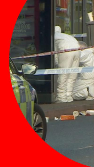 The front right tyre of a police car in front of some crime scene tape. Behind the tape, two forensics officers in white suits are carrying out an investigation.