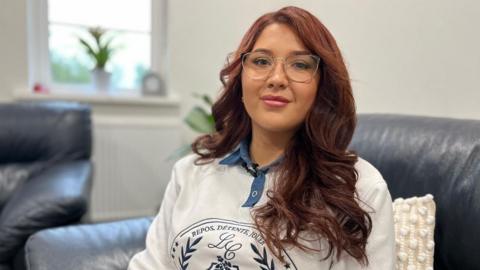 Megan has long, wavy reddish brown hair. She is wearing glasses, a white and blue top, and is smiling at the camera. She is sat on a dark leather chair, with a window and white walls in the background