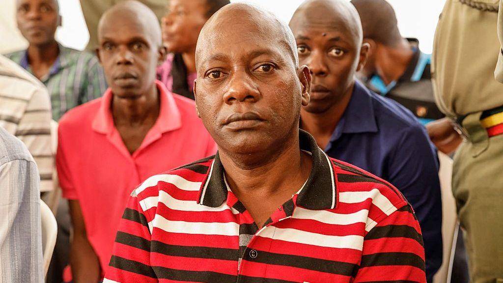 Paul Mackenzie, wearing a red, black and white striped shirt, sits in court and looks at the camera.