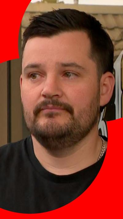 A man dressed in a black T-shirt with a beard and brown hair standing out the front of his barber shop.