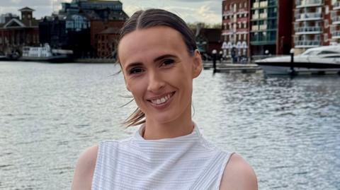 A woman with brown hair that is tied up in a ponytail smiling, she is wearing a white sleeveless top, the background is a dock.