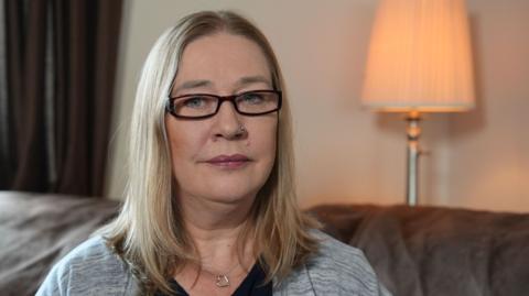 Valerie Stobbart has shoulder-length blonde hair and narrow, black-rimmed glasses and has a neutral expression. She is wearing a grey cardigan and black top, with a silver necklace with a charm in the shape of an outline of a heart on it. She is sitting on a dark sofa and behind her are brown curtains and a cream lamp-shaded standard lamp against a cream wall. The lamp is switched on, emitting a warm peach glow. 