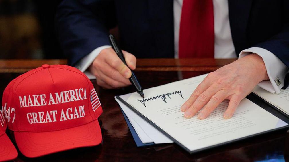 US President Donald Trump signs an executive order in the Oval Office at the White House in Washington, DC on 23 April 2025.  A red baseball hat embroidered with "Make America Great Again" in white thread sits to the left of the document, which shows Trump's distinctive signature